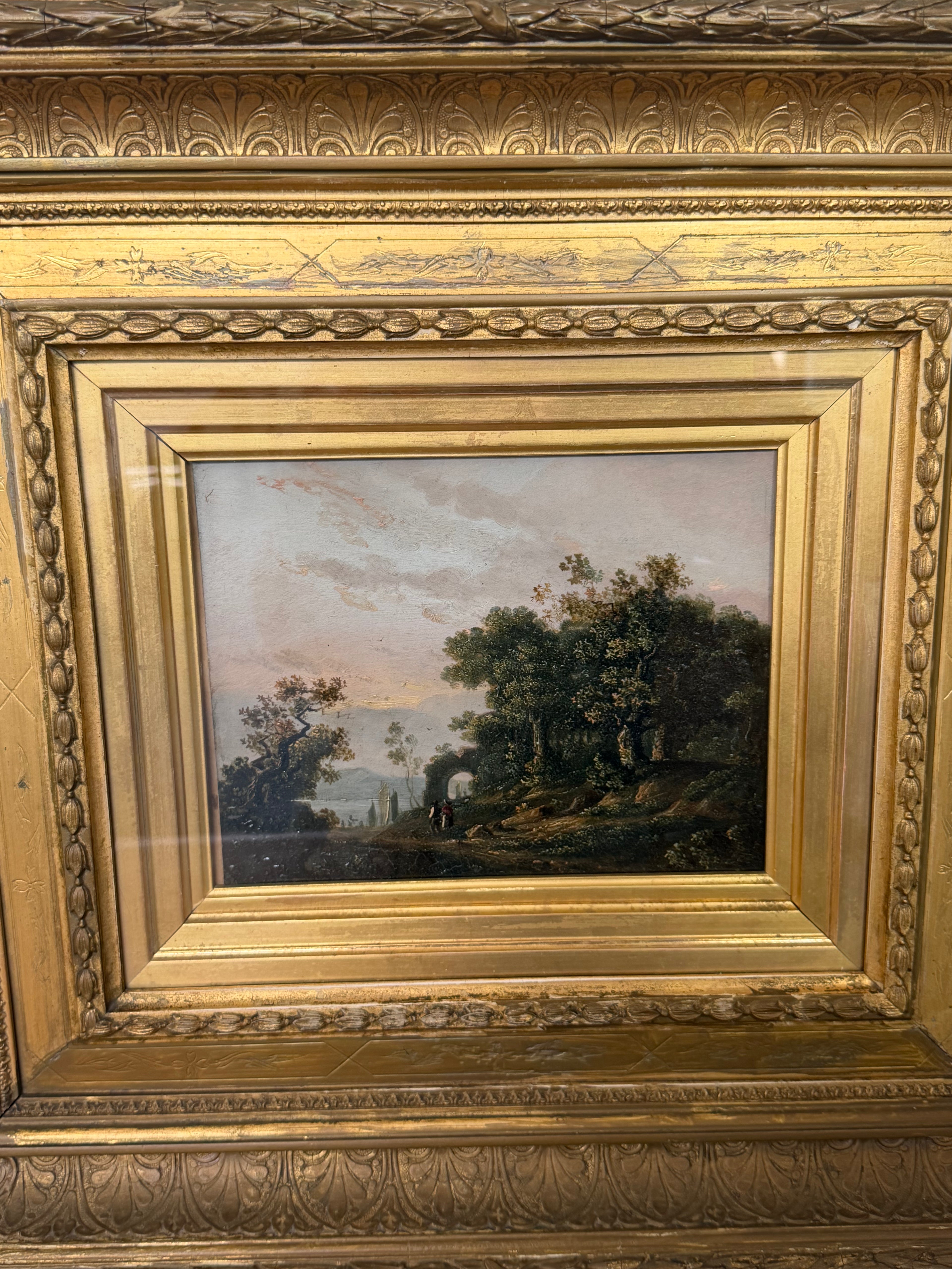 An antique landscape painting housed in an ornate, carved gold frame. The painting depicts a scenic forest path with trees and a distant figure under a cloudy sky, while the frame features intricate classical detailing.